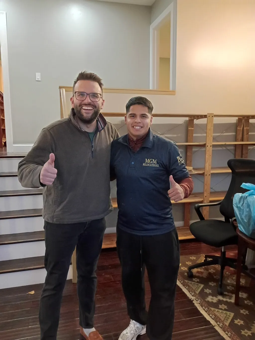 Two men, one in an MGM Relocations shirt, smiling and giving thumbs up indoors.
