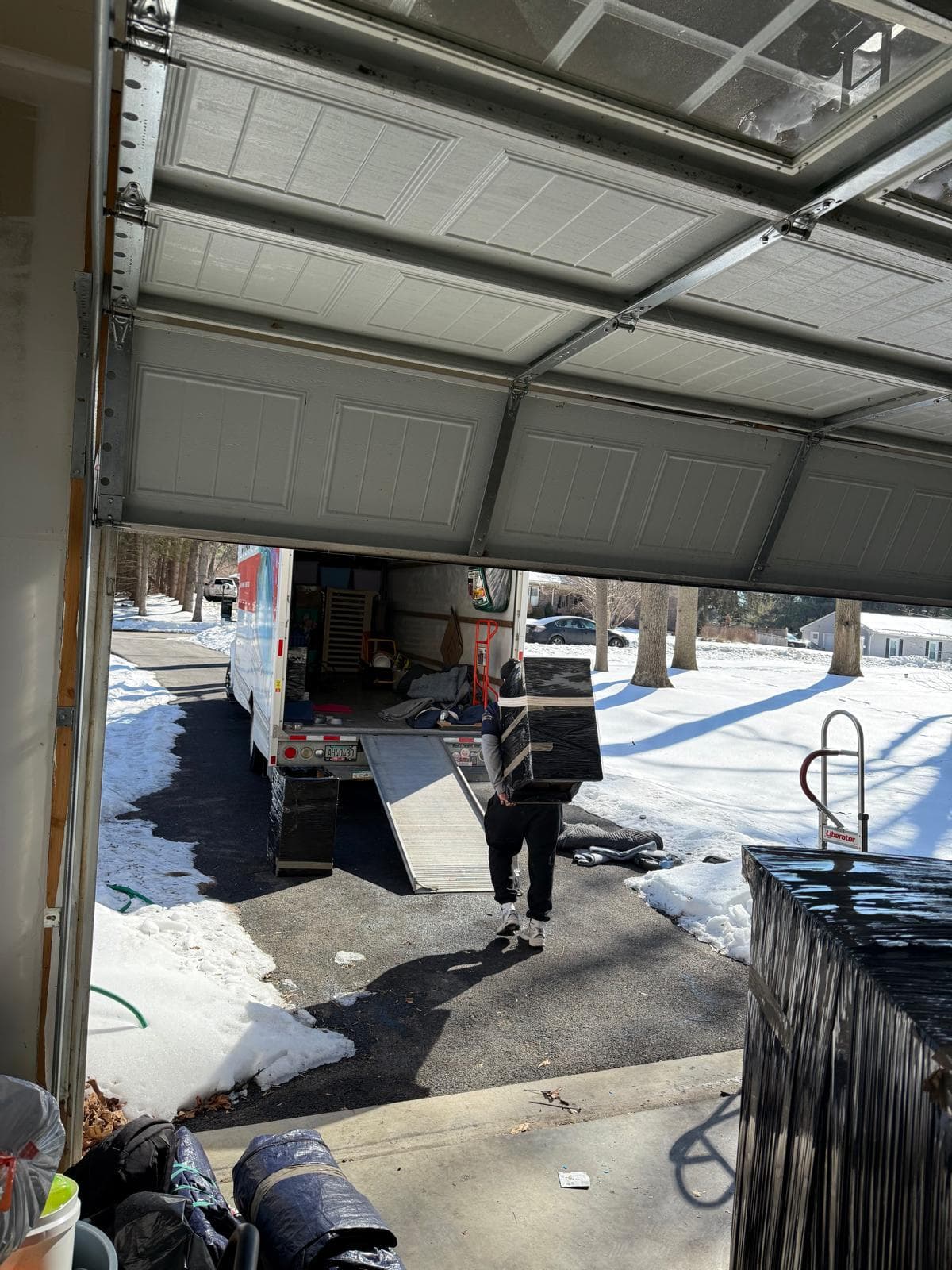 Person carrying a large wrapped box toward a moving truck in a snowy driveway.