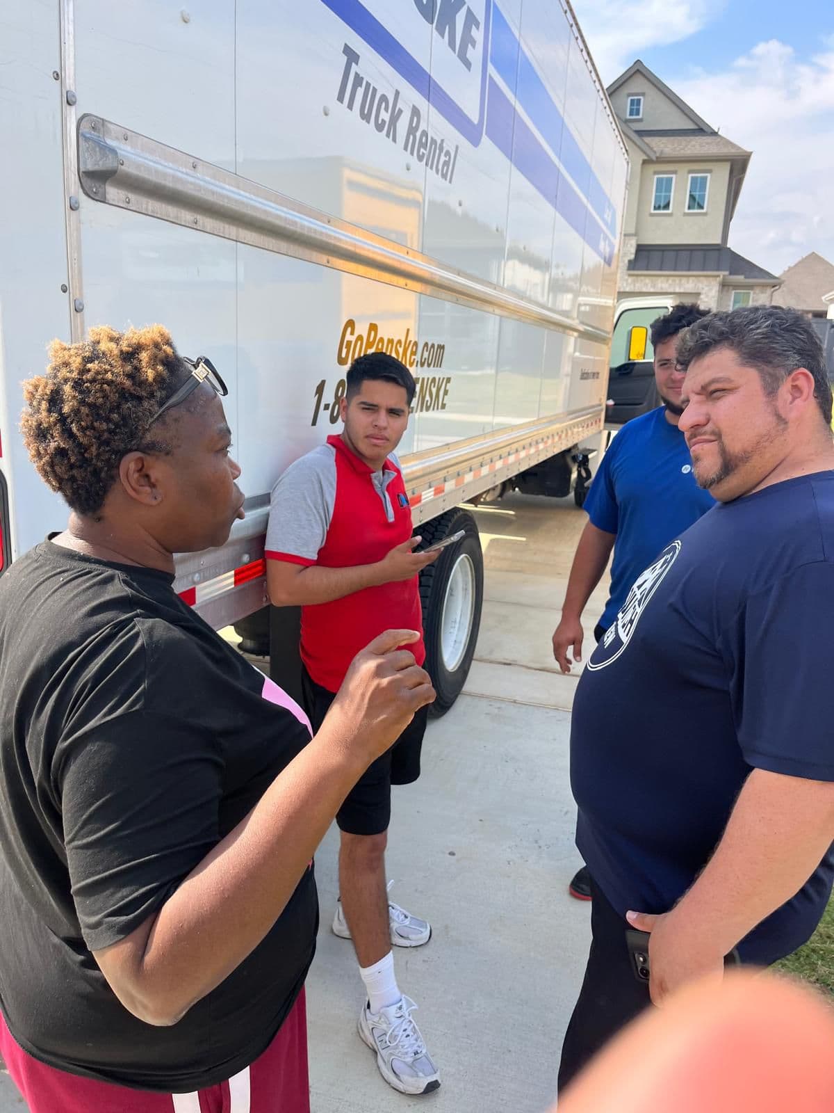 A woman gestures while talking to three men beside a white Penske rental truck.