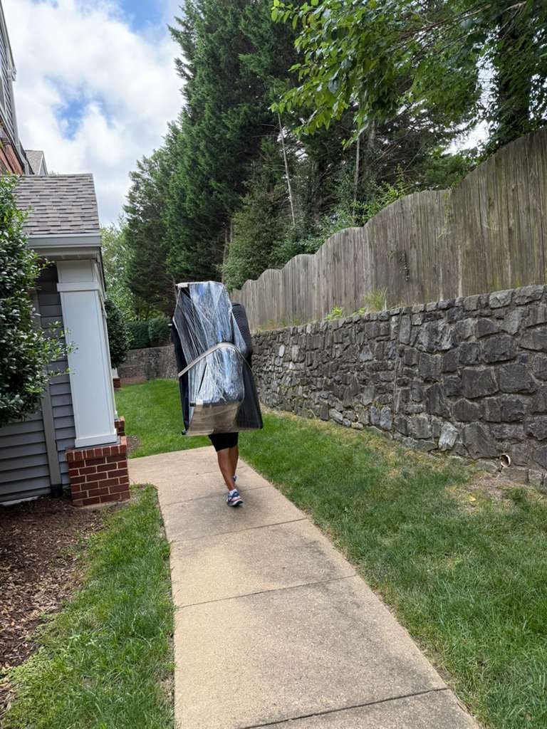 Person carrying a large, plastic-wrapped object on their back along a concrete sidewalk.