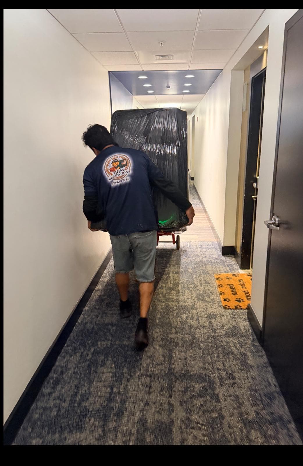 A professional mover pushes a large item wrapped in black plastic down a carpeted hallway.